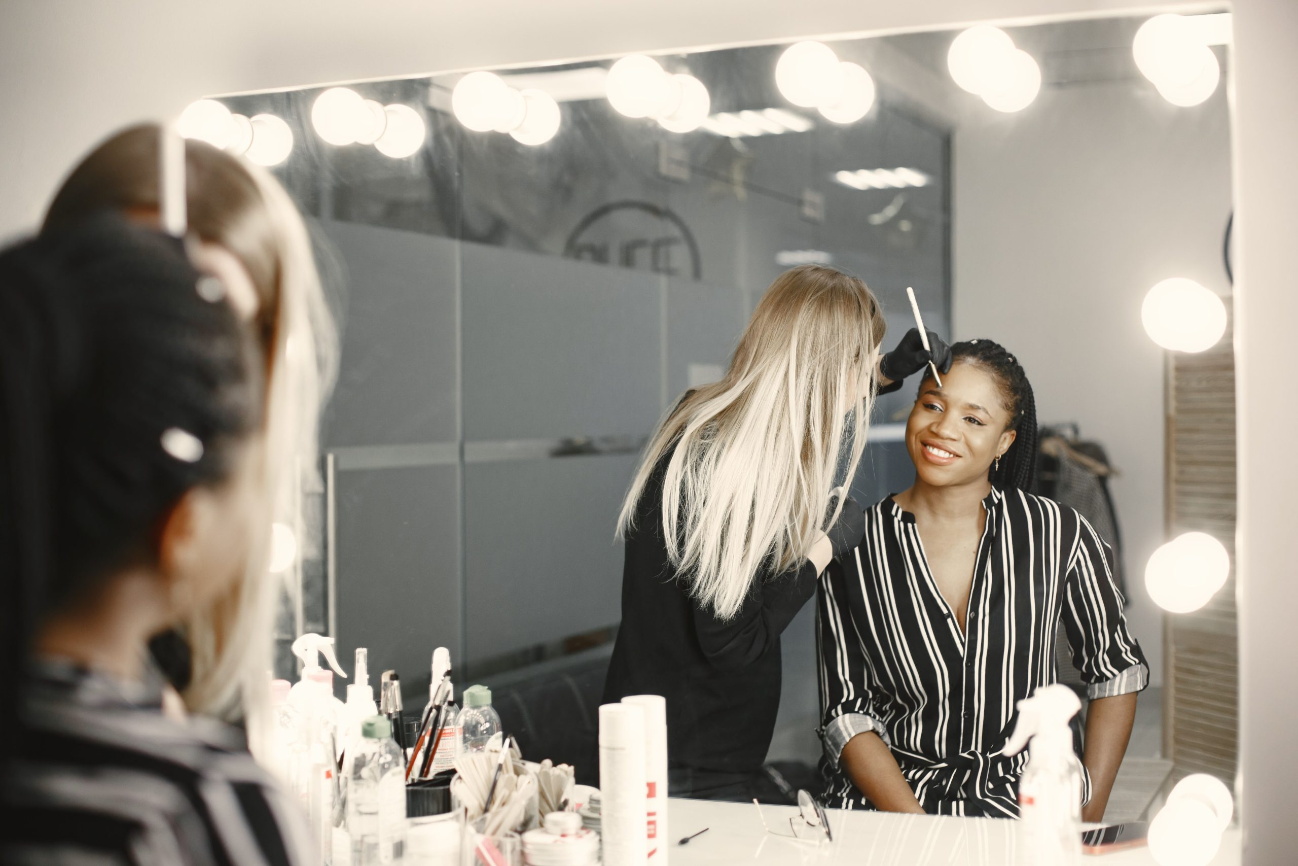 A professional makeup artist applies makeup to a smiling woman in a well-lit backstage setting.